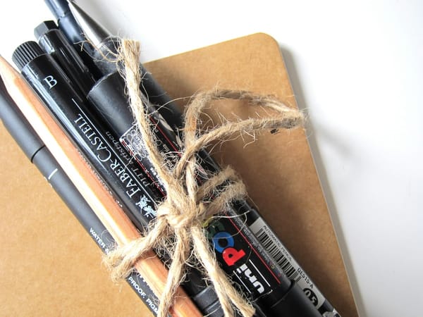 A bunch of pens and pencils are tied together with twine and lying on a notebook with a kraft paper cover.